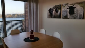 Uma área de refeições com uma mesa redonda, cadeiras e um objeto decorativo. Há duas pinturas na parede retratando animais. Uma janela com vista para uma paisagem montanhosa é visível.