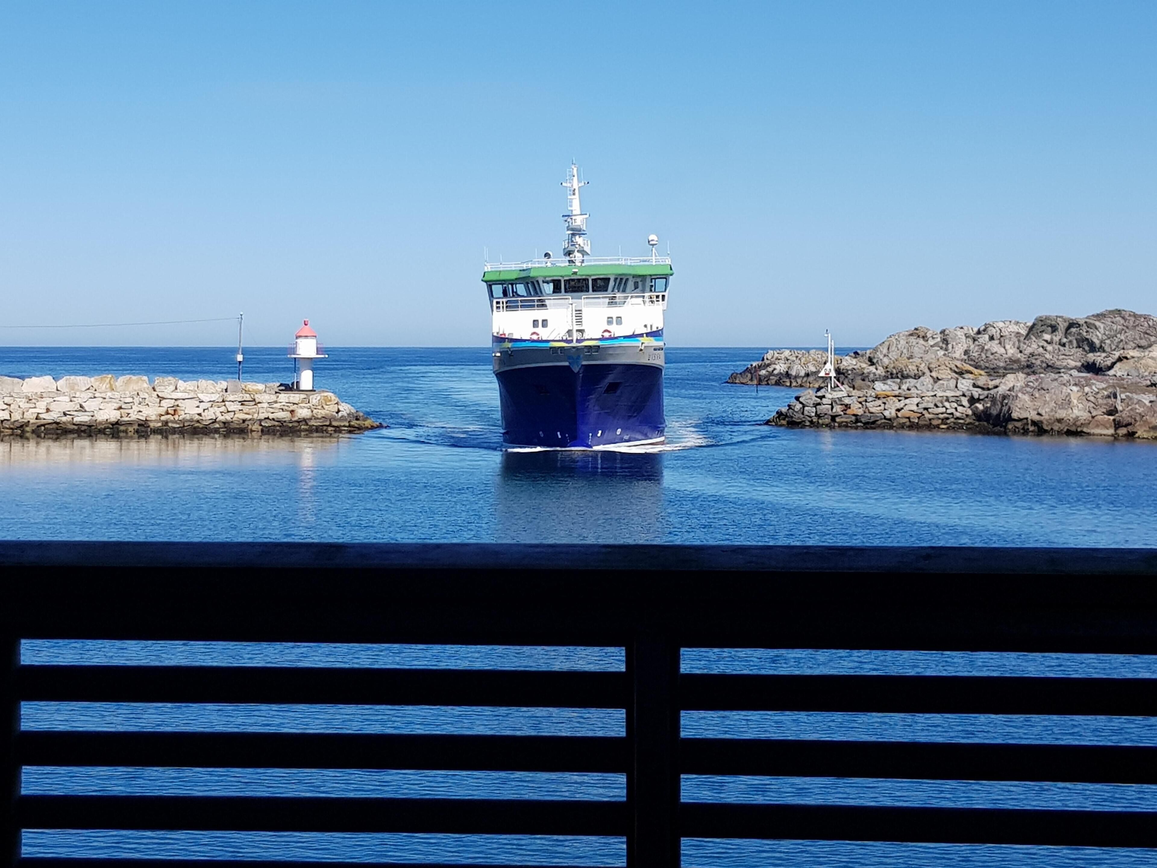 A large ship docked at a harbor with a lighthouse and rocky outcrops in the background.