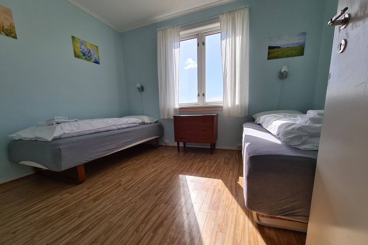 Una habitación con dos camas individuales, suelo de madera, una cómoda de madera pequeña y una ventana con cortinas.