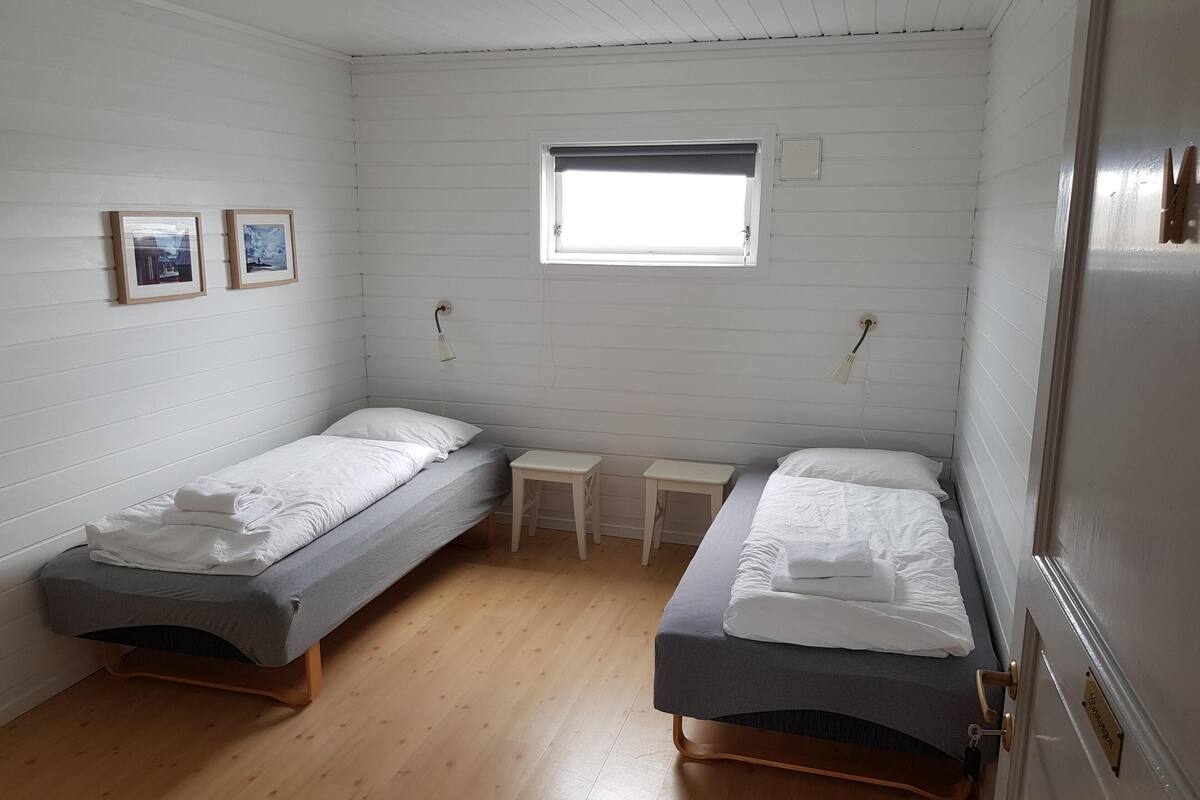 Una habitación pequeña y limpia con dos camas individuales, una mesita y una ventana.