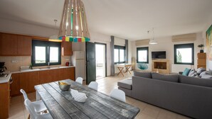 A modern kitchen with wooden cabinets, a dining table with chairs, a gray sofa, and a TV in the living area. - Portela Villas Kapsali (Viannos)
