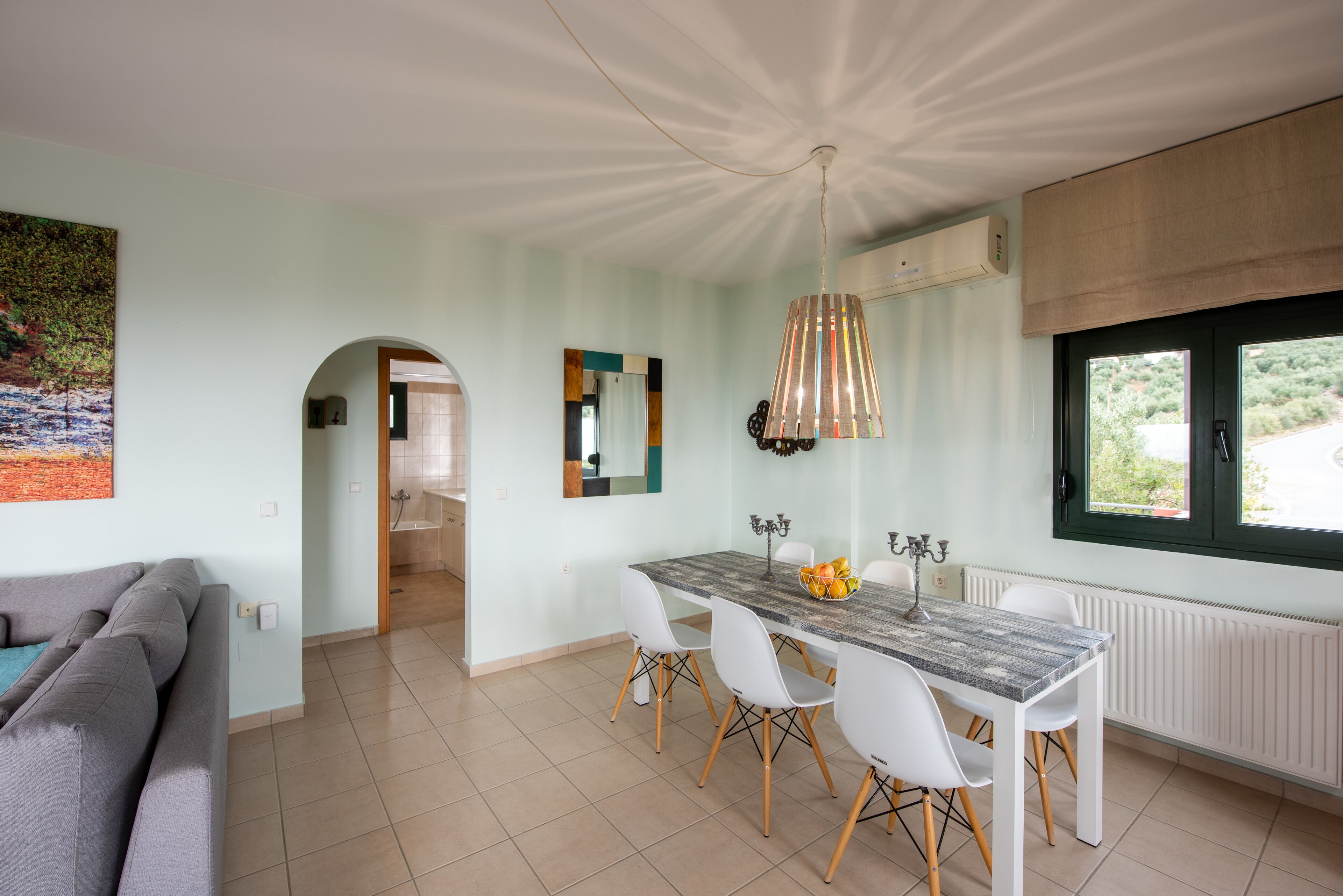 A modern kitchen with a dining table, chairs, and a pendant light. There is a window with a view of greenery outside.