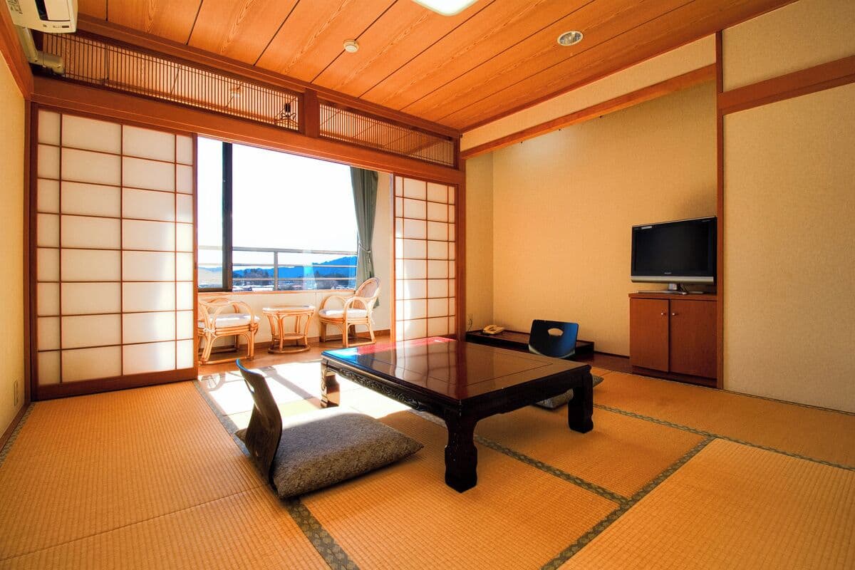 Une pièce de style japonais traditionnel, avec un sol recouvert de tatamis, une table basse, un téléviseur et une vue sur l’extérieur.