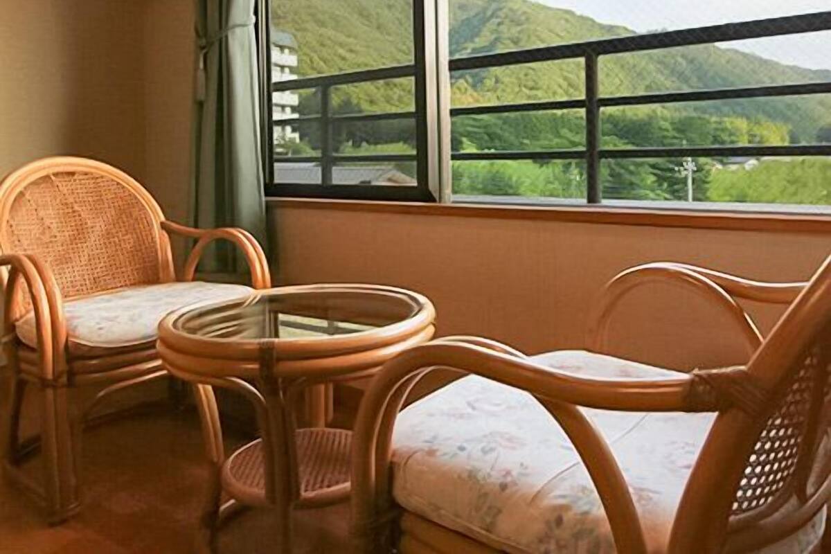 Ein Zimmer mit zwei Korbstühlen und einem kleinen Tisch, ein großes Fenster mit Blick auf grüne Hügel.