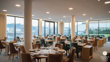 Ampla área de jantar de hotel, com várias mesas e cadeiras, e janelas grandes com vista para a cidade.