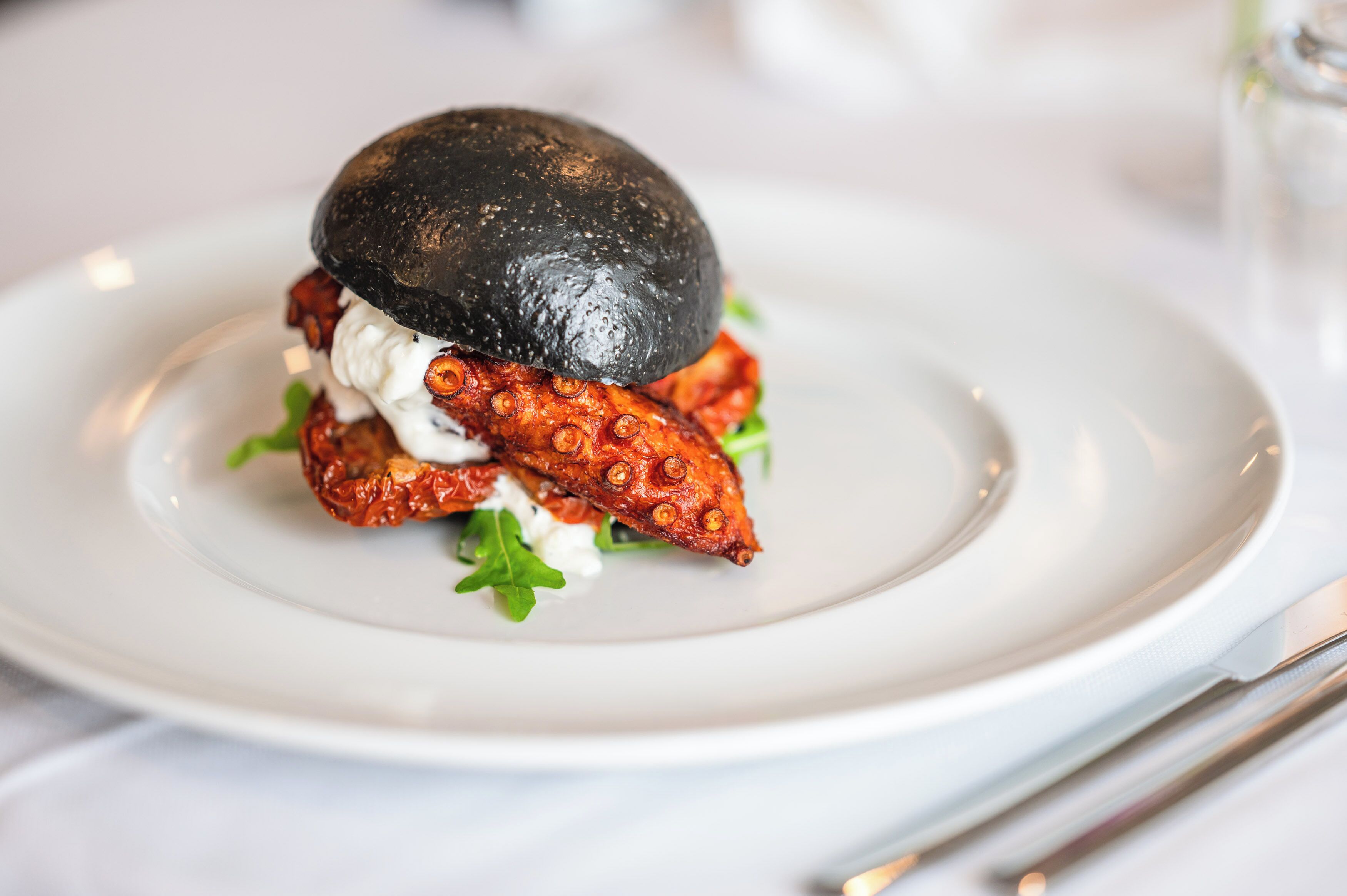 Une assiette avec un hamburger et un pain noir, garni de légumes verts et d’une sauce crémeuse.