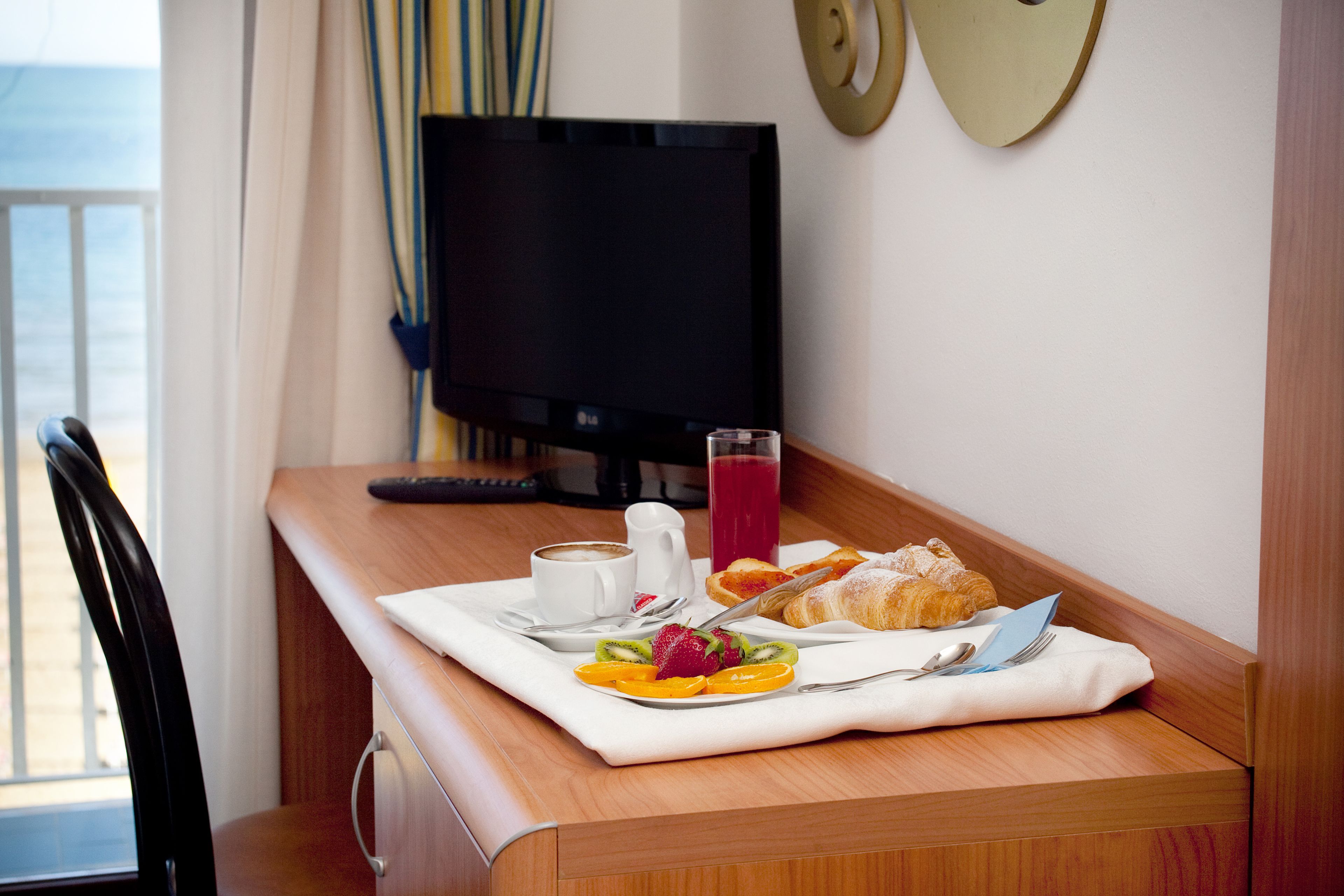 A hotel room with a wooden desk, a television, breakfast items including croissants, fruit, and a glass of juice.