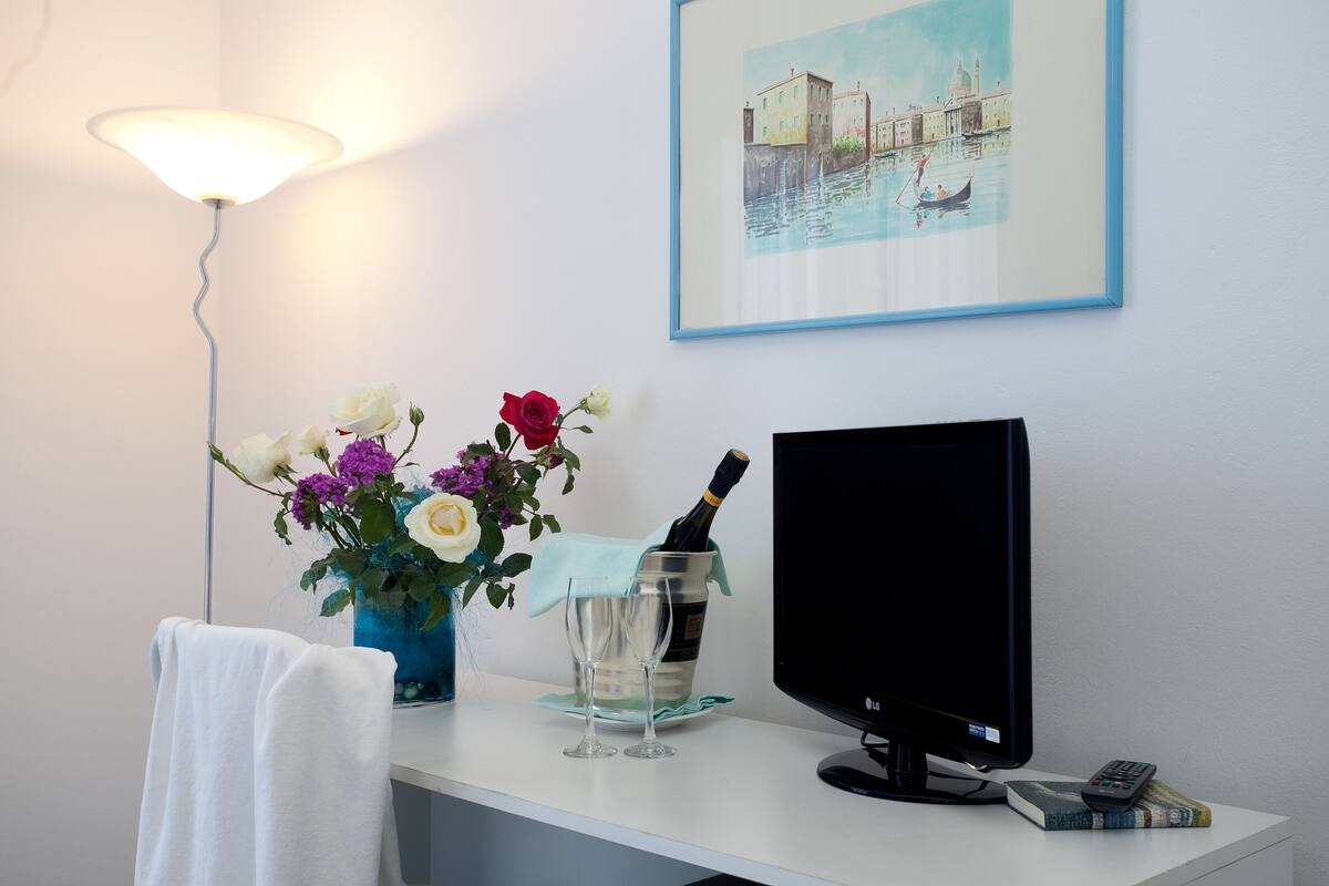 A hotel room with a white desk, a TV, a remote control, a vase with flowers, a bottle of champagne, and a framed picture on the wall.