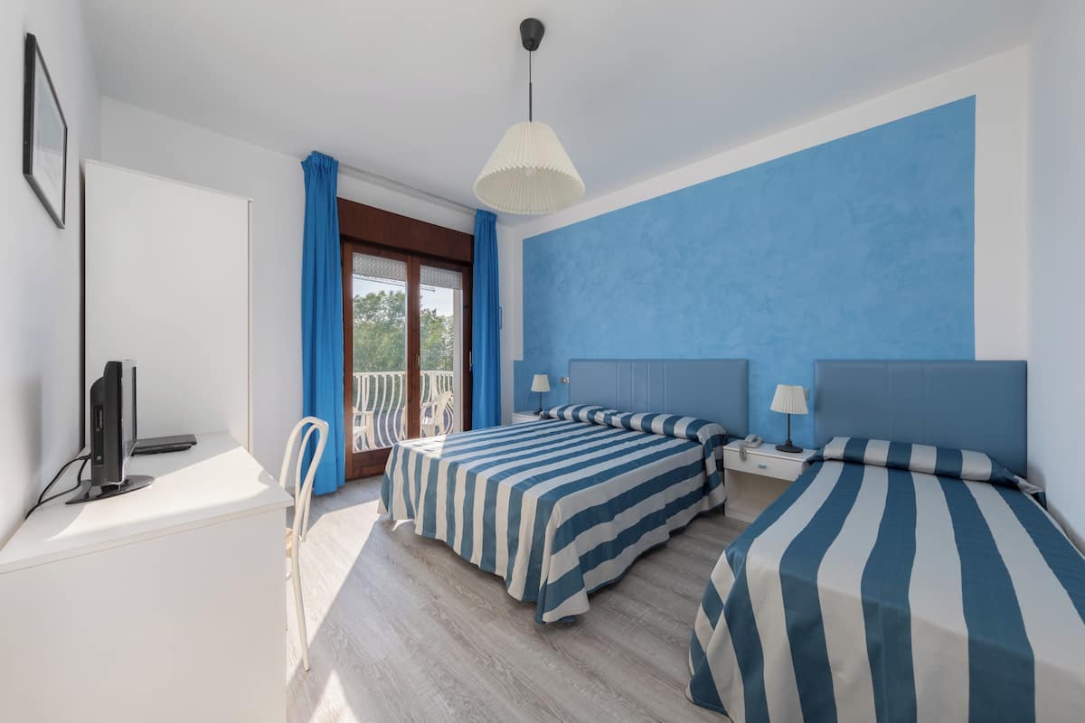 A hotel room with two beds, a desk with a computer, a chair, and a balcony with blue curtains.