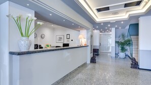 A modern reception area with a marble counter, a vase of white flowers, a clock, framed artwork, and a lamp. - Hotel Gritti (Jesolo)