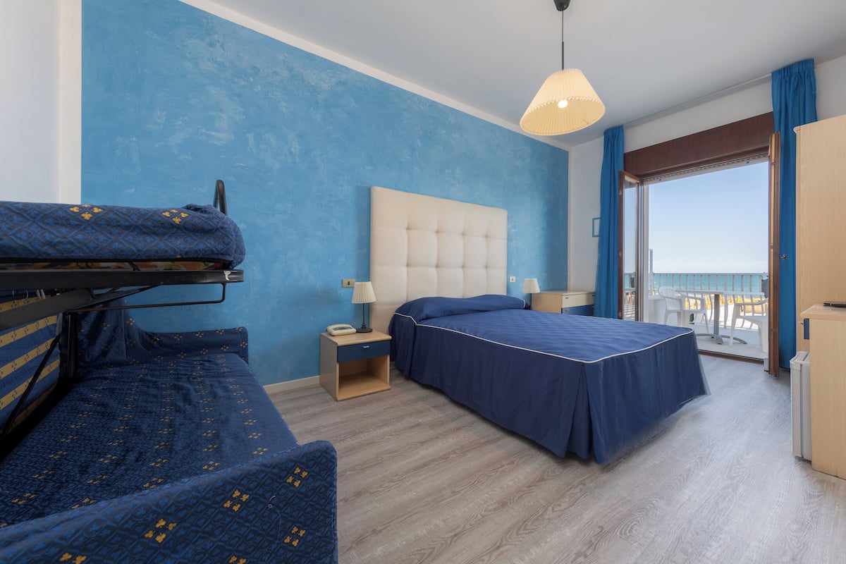 A hotel room with a bunk bed, a single bed, a nightstand with a lamp, a large window with a view of the sea, and a balcony with outdoor furniture.