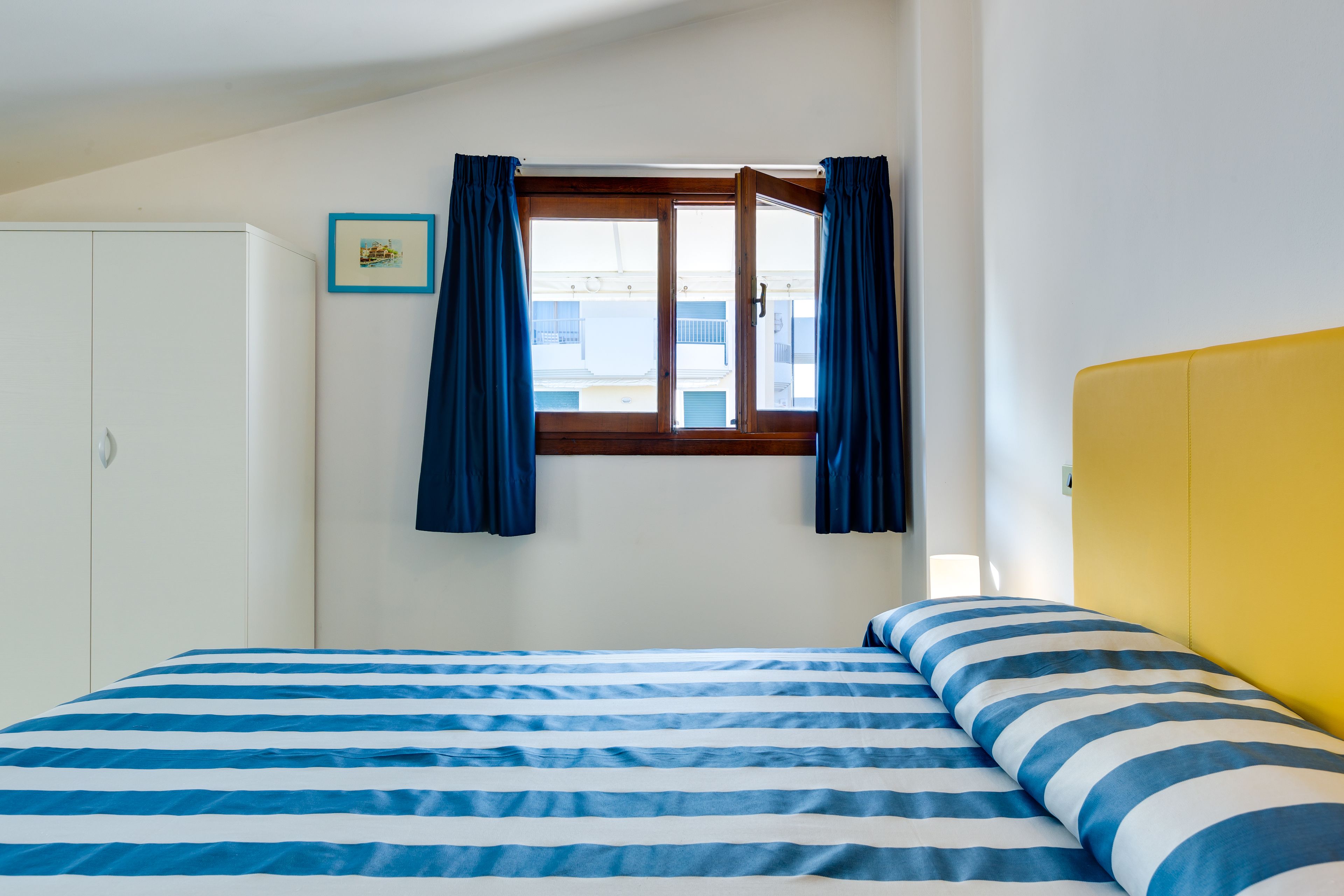 A bedroom with a bed, a window with curtains, a white wardrobe, and a yellow headboard.