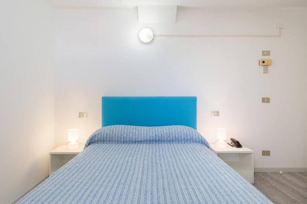 A bedroom with a blue headboard, a white nightstand with a lamp, and a bedside table with a phone.