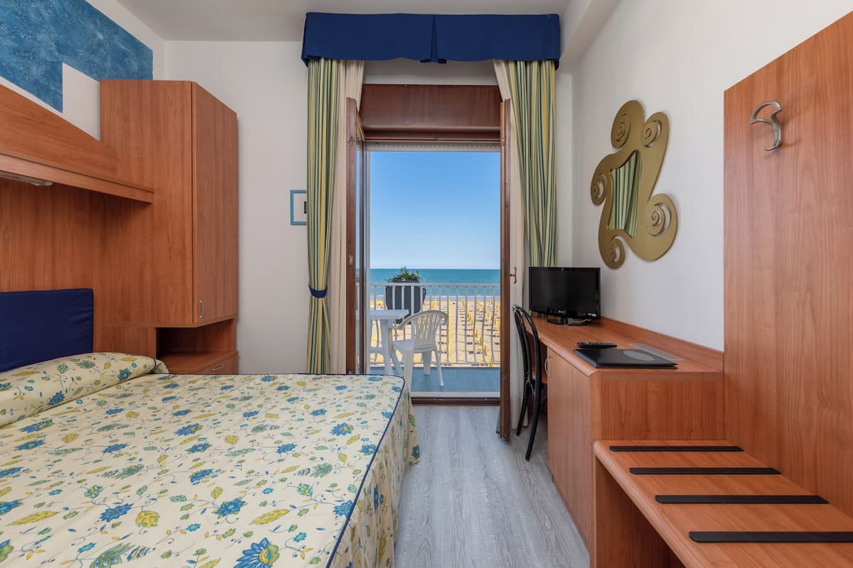 A hotel room with a bed, a desk, a TV, and a balcony with a view of the sea.
