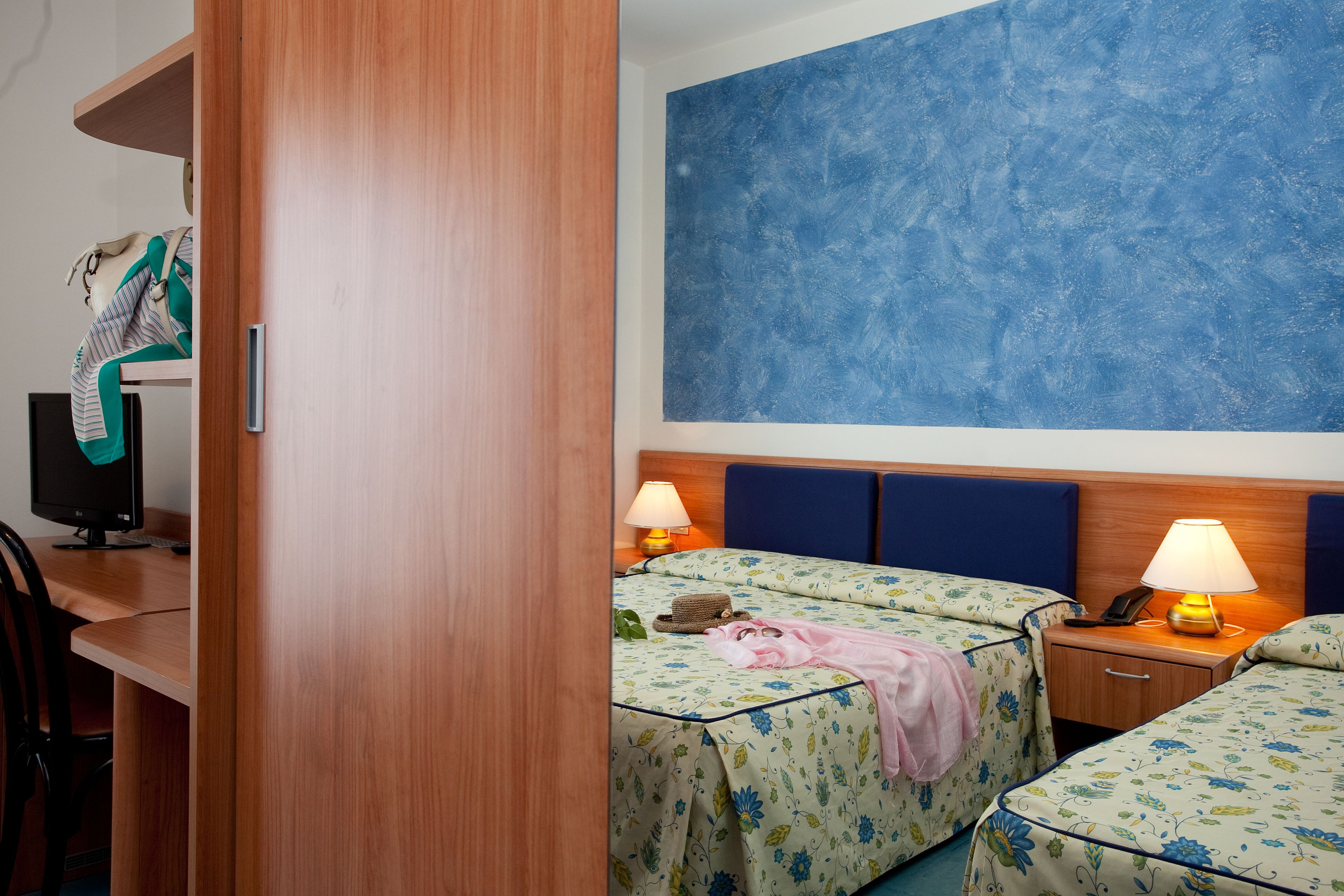 A hotel room with two beds, a desk with a computer, and a large blue wall.