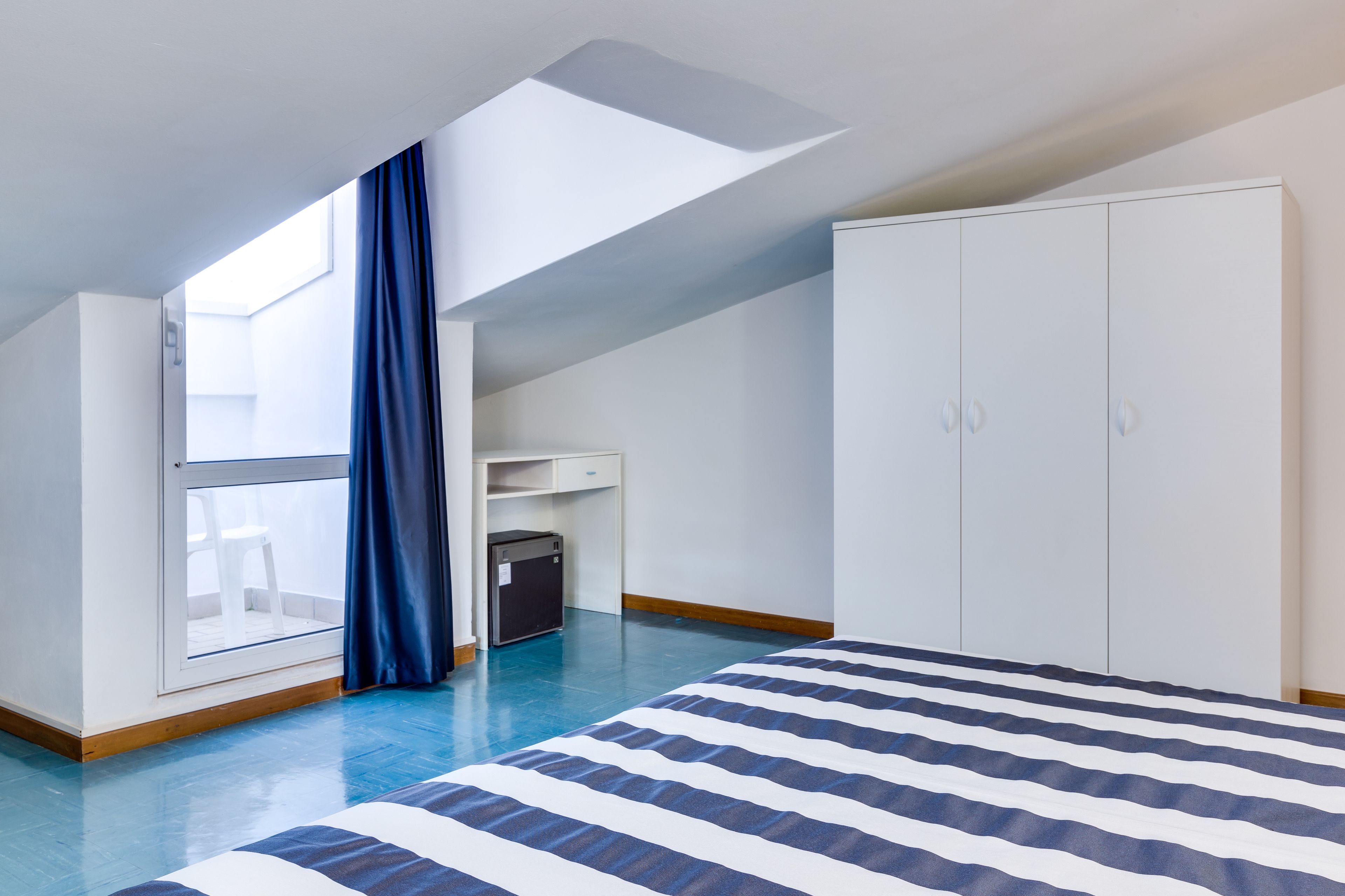 A bedroom with a skylight, a bed with striped bedding, a wardrobe, and a small desk area.