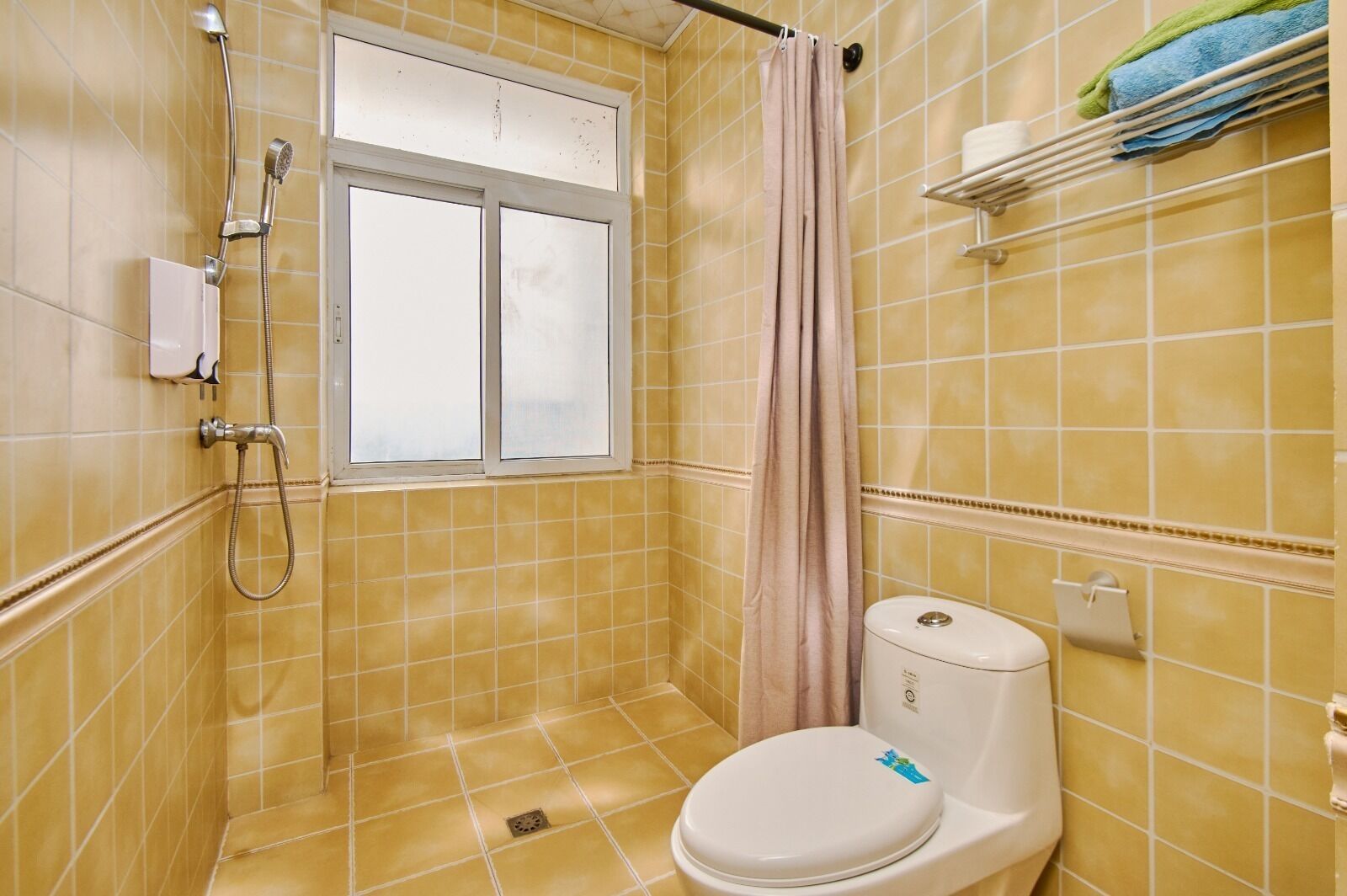 A bathroom with yellow tiles, a window, a shower, a toilet, and a towel rack with towels.