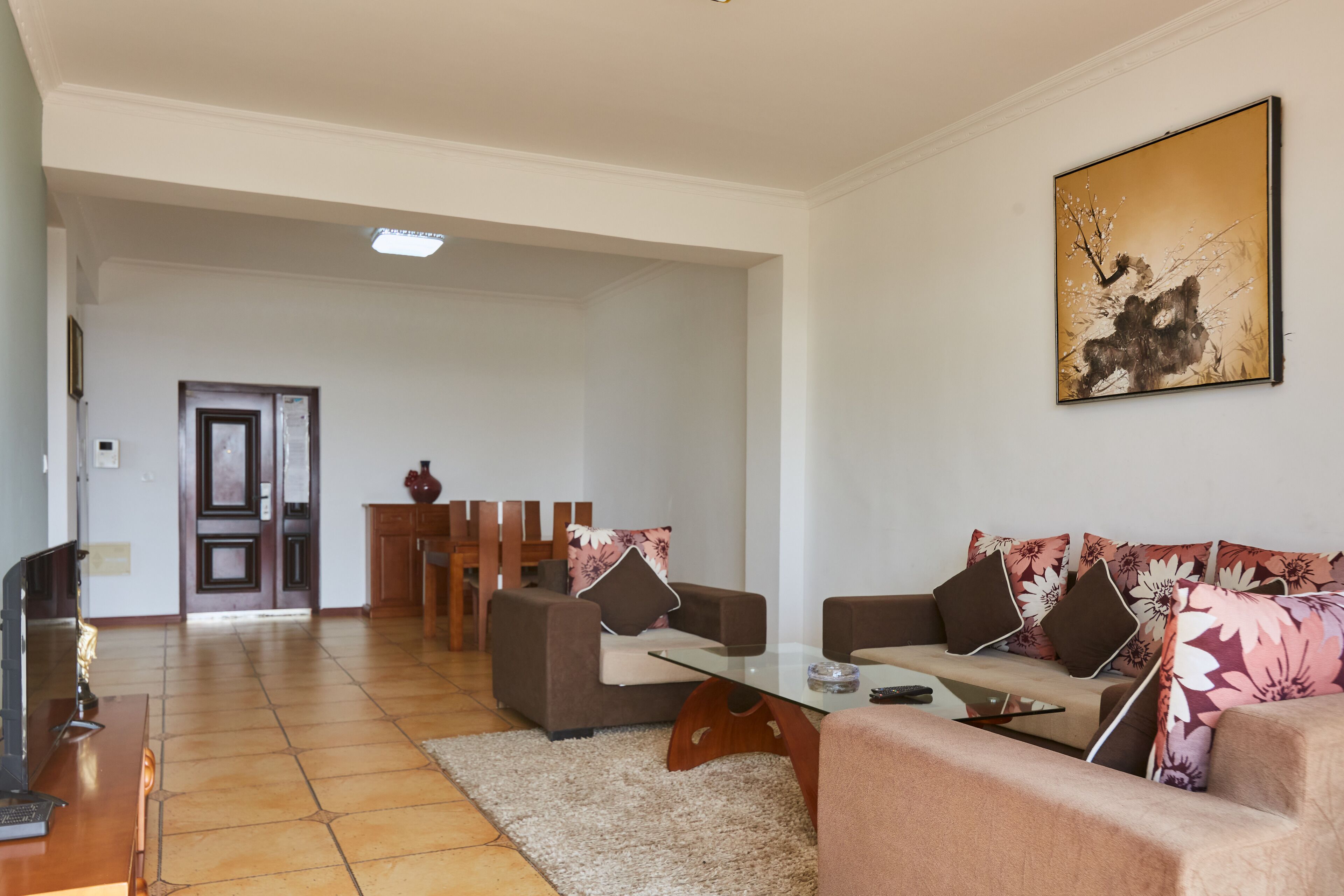 A spacious living room with a sofa set, a coffee table, and a TV. There is a door leading to another room and a painting on the wall.