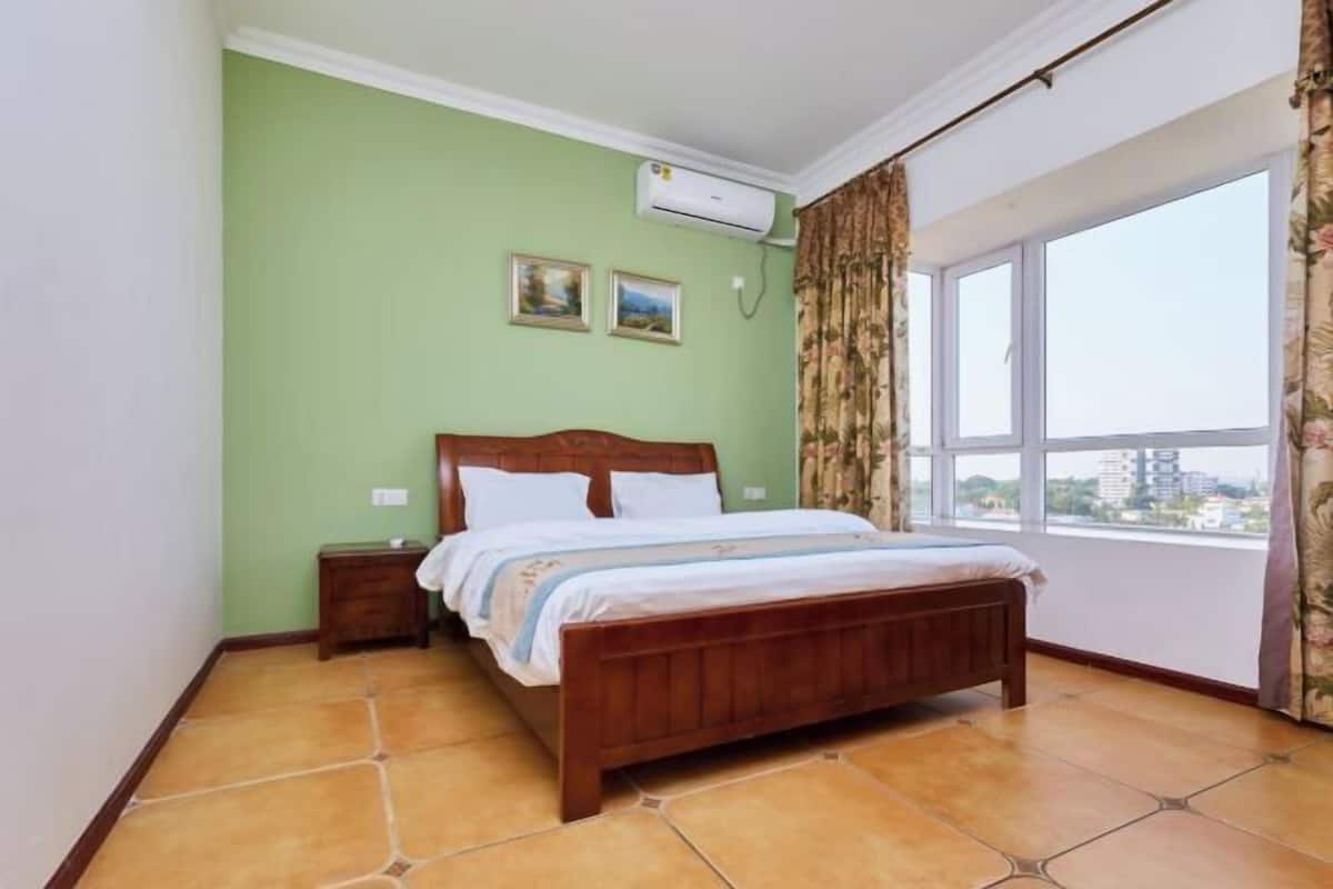 Une chambre à coucher comprenant un lit, une table de chevet, un appareil de climatisation, deux tableaux encadrés et une fenêtre avec des rideaux.