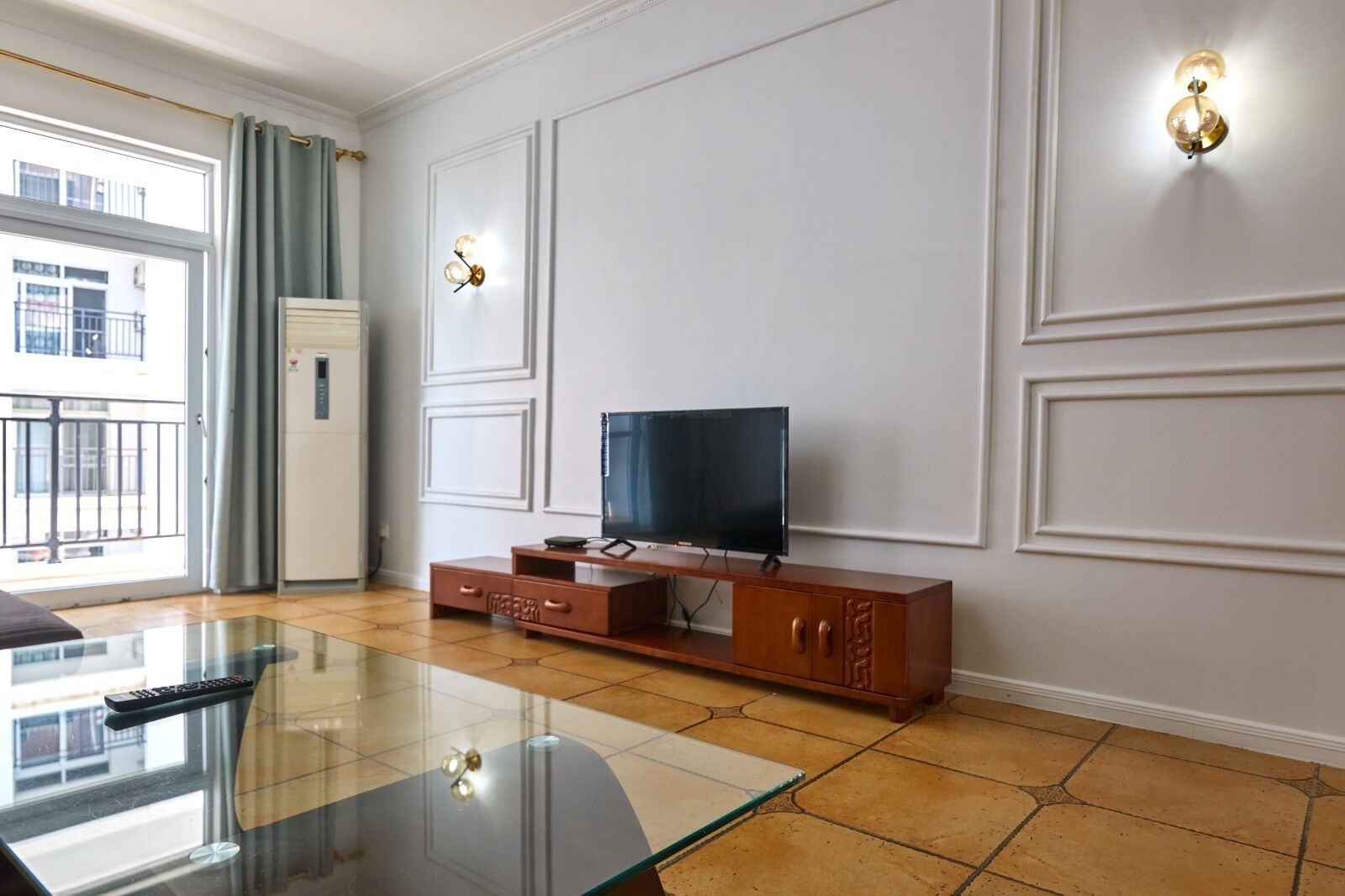 Una sala de estar moderna con una mesa de centro de vidrio, un televisor de pantalla plana sobre un mueble de madera, y un balcón con vistas a la ciudad.
