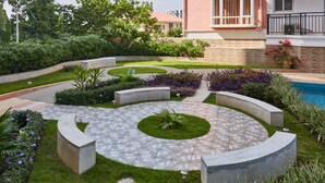 Un jardí paisatjistic amb un pati circular de pedra, una piscina i un edifici amb un balcó.