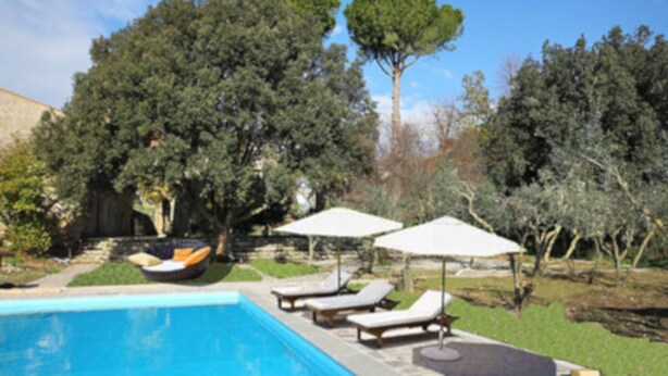 Una piscina con sillones de descanso y sombrillas, rodeada de árboles y un muro de piedra.
