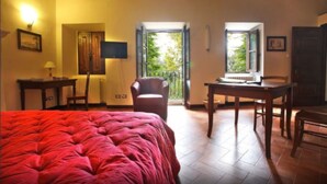 Una habitación con una cama grande, un escritorio, una silla y una mesa. Hay dos ventanas con contraventanas abiertas y una puerta que da a otra habitación.
