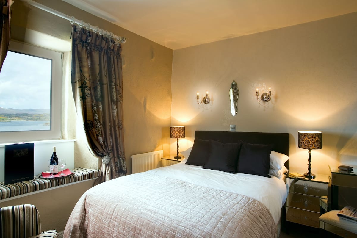 Een hotelkamer met een groot bed, twee bedlampjes, een raam met gordijnen, uitzicht op het landschap en een klein tafeltje met een fles en glazen.