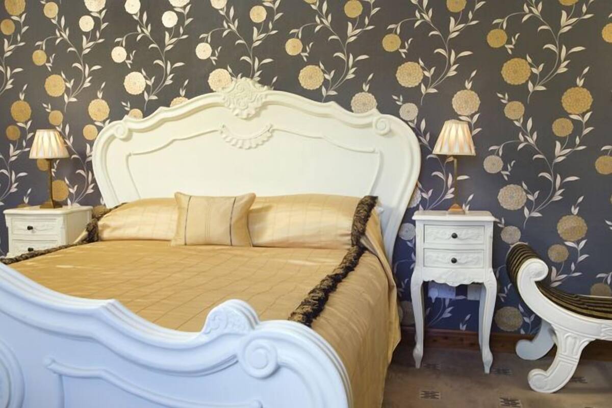 Een slaapkamer met een wit, versierd bed, twee nachtkastjes met lampen, en behang met een patroon.