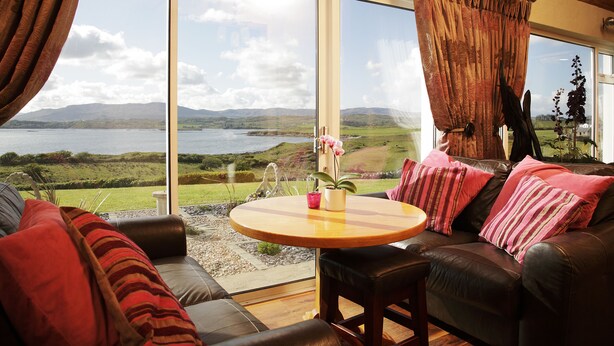 A living room with a large window offering a scenic view of a lake and hills. There is a leather sofa, a round wooden table, and a potted plant. - Castle Murray House Boutique B&B (Dunkineely)