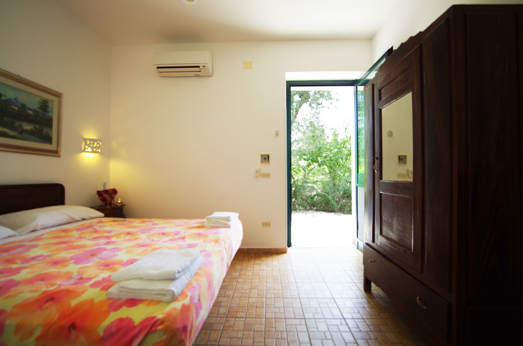 A bedroom with a bed, an air conditioner, a nightstand, a painting, and a view of the outdoors.