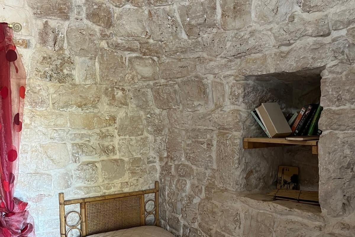 Una habitación con pared de piedra, cama con cabecera de madera, estante con libros y cortina roja.