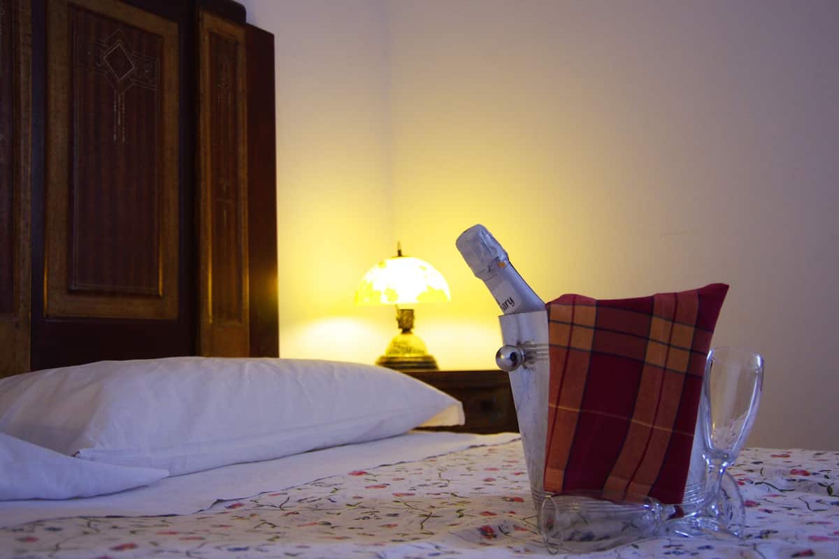 Una cama con un almohadón blanco y una colcha estampada con flores. Una botella de vidrio con un paño a cuadros rojo y marrón está colocada sobre la cama. También se ve un vaso de vidrio en la cama.