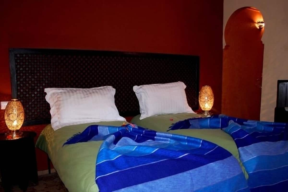 A hotel room with a large bed, two pillows, a nightstand with a lamp, and a wall-mounted light fixture.