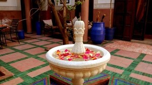 Uma fonte com água e pétalas de flores num pátio com azulejos estampados e plantas em vasos.