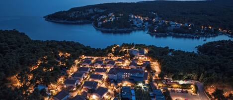 Vista aérea de un pueblo costero al anochecer, con edificios iluminados, un puerto deportivo y una colina boscosa en el fondo.