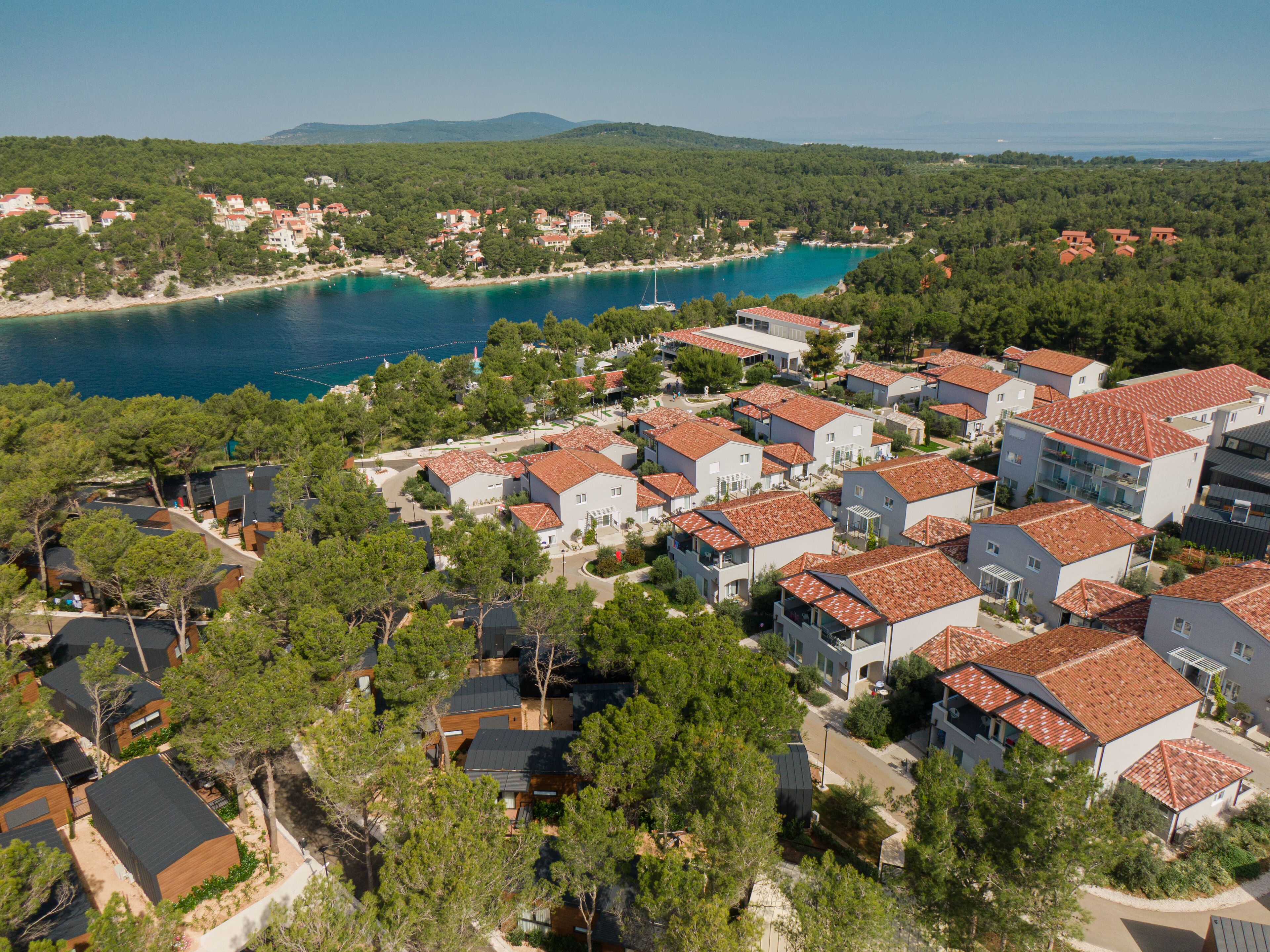 A coastal residential area with houses featuring red-tiled roofs, surrounded by greenery and a clear blue sea.