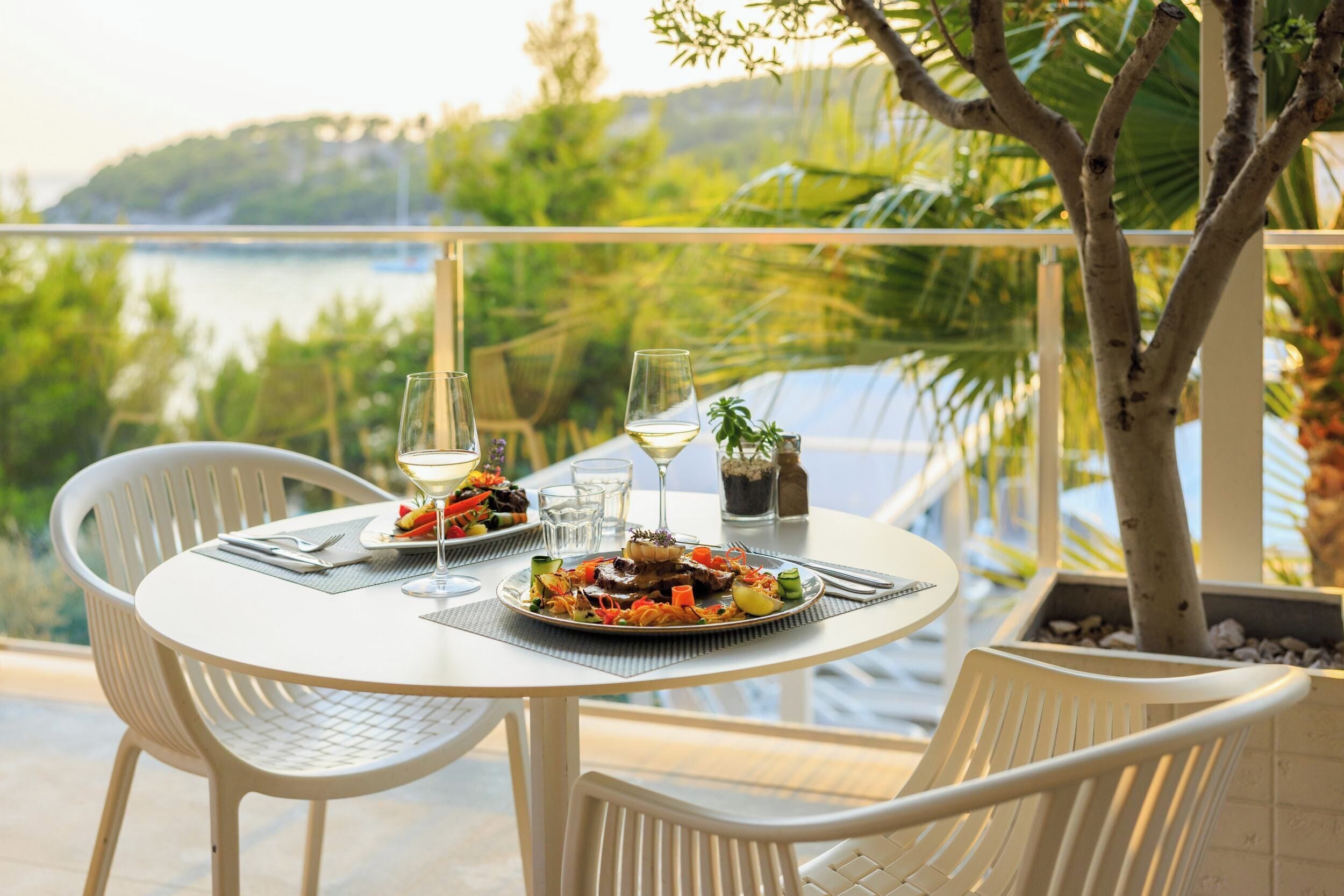 A table set with plates of food, wine glasses, and a bottle of water, overlooking a scenic view of a body of water and greenery.