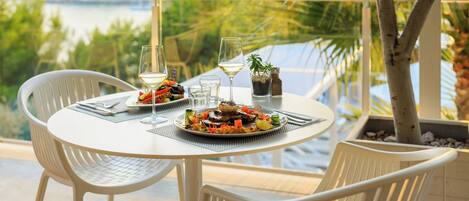 A table set with plates of food, wine glasses, and a bottle of water, overlooking a scenic view of a body of water and greenery.