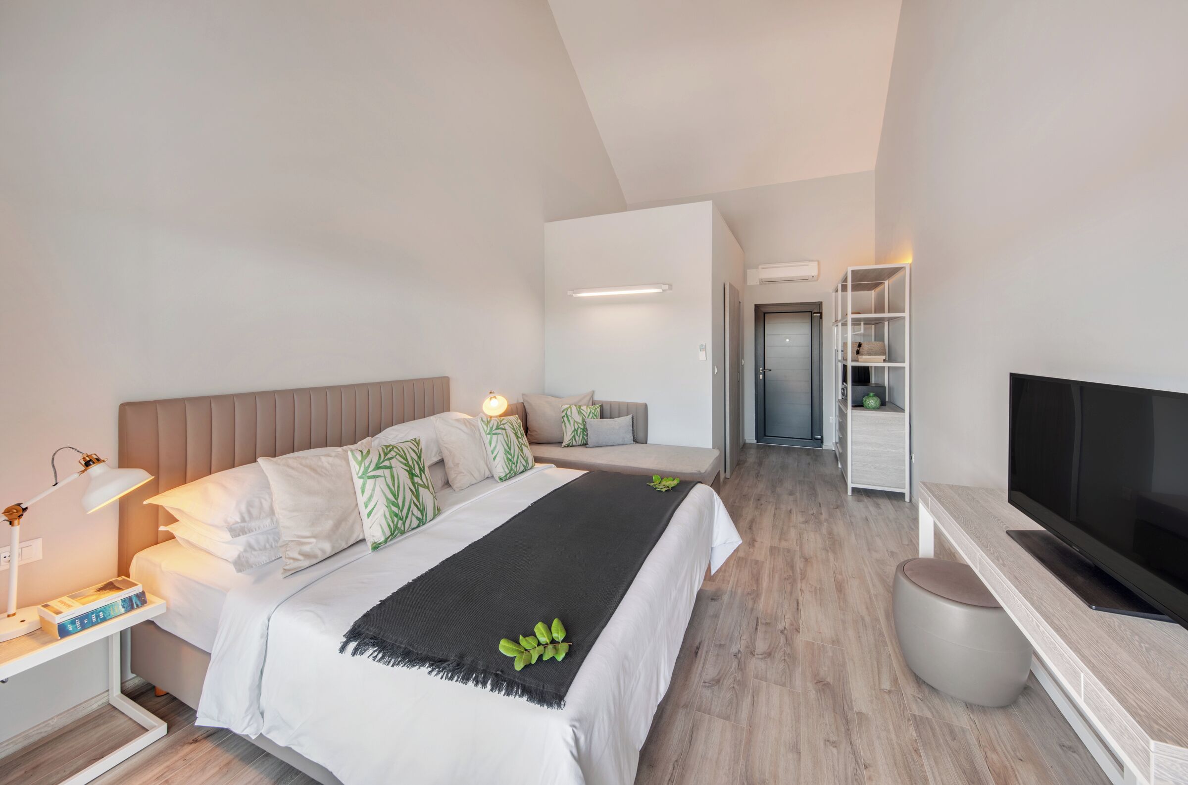 A modern hotel room with a bed, bedside table with a lamp, a TV, a wardrobe, and a small seating area.