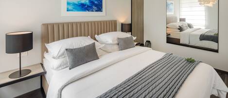A modern bedroom with a large bed, a bedside table with a lamp, a wall-mounted mirror, and a framed picture.