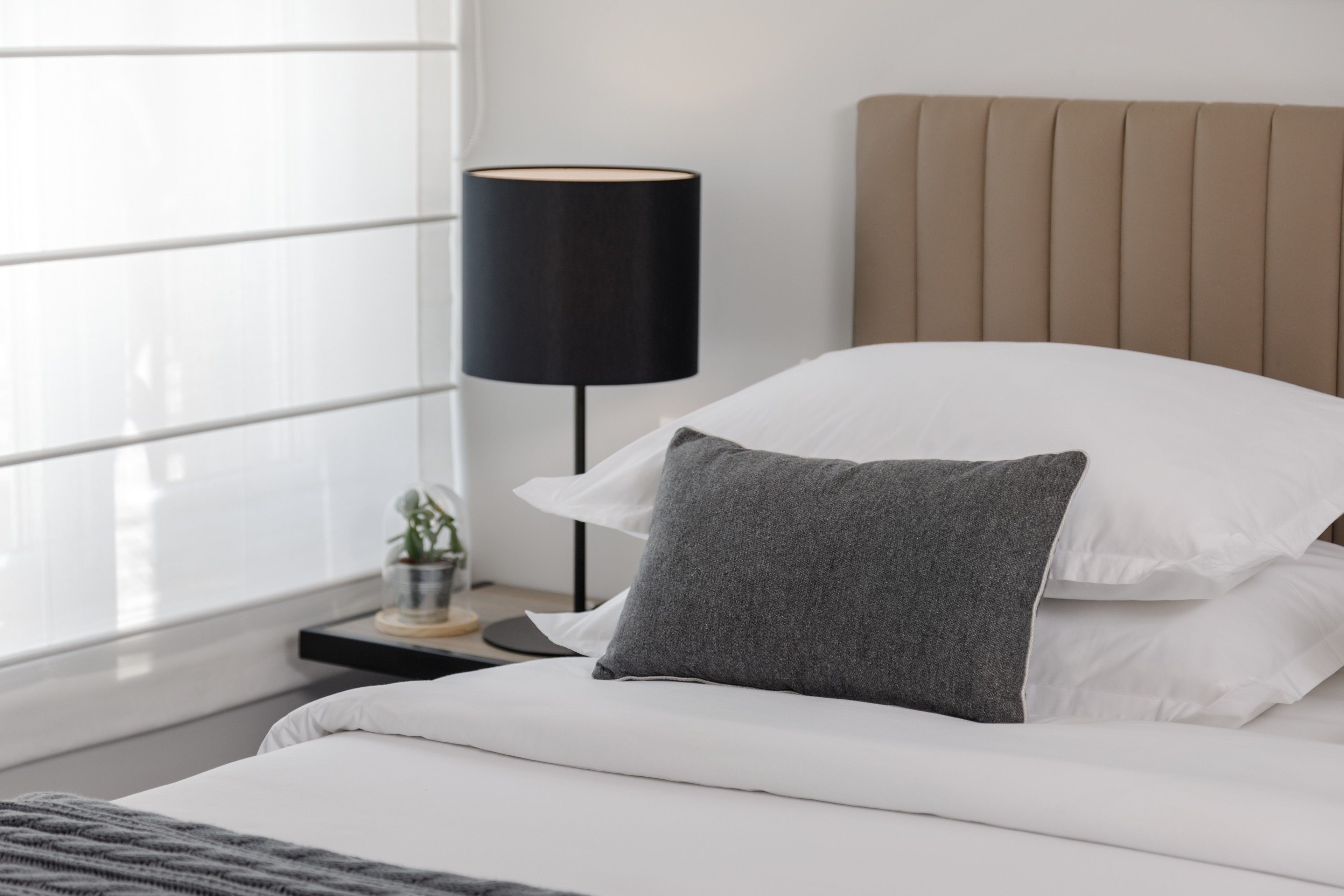 Ein ordentlich bezogenes Bett mit weißer und grauer Bettwäsche, eine schwarze Stehlampe und ein beige farbener Kopfteil.