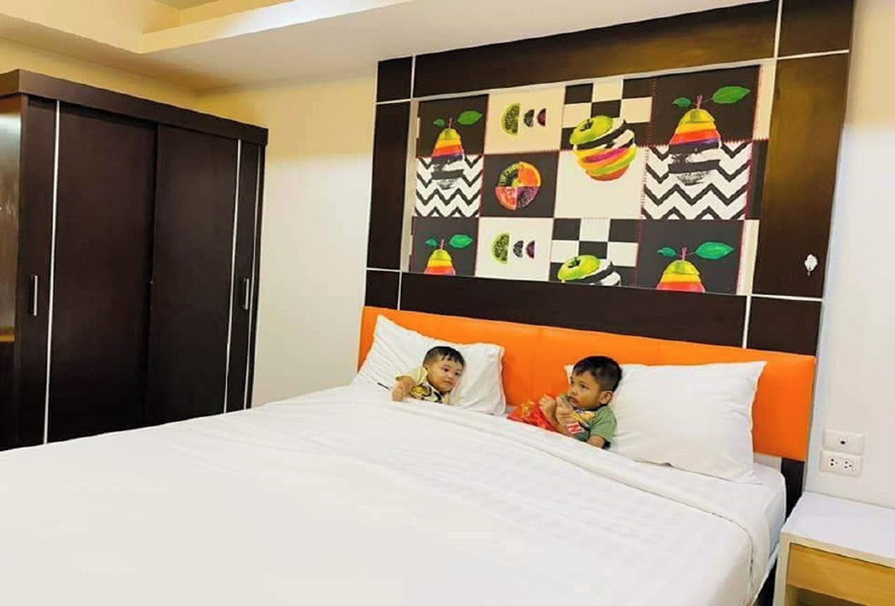A bedroom with a bed, two pillows, a nightstand, and a wall with a colorful fruit pattern.