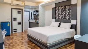 A modern bedroom with a large bed, a built-in desk, and a kitchenette in the background. - The Ring Residence (Hat Yai)