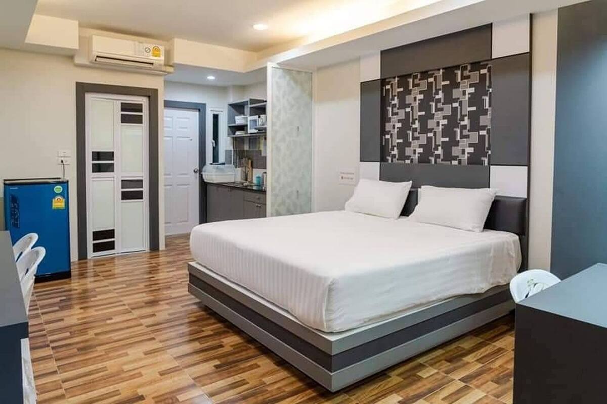 A modern bedroom with a large bed, a built-in desk, and a kitchenette in the background.
