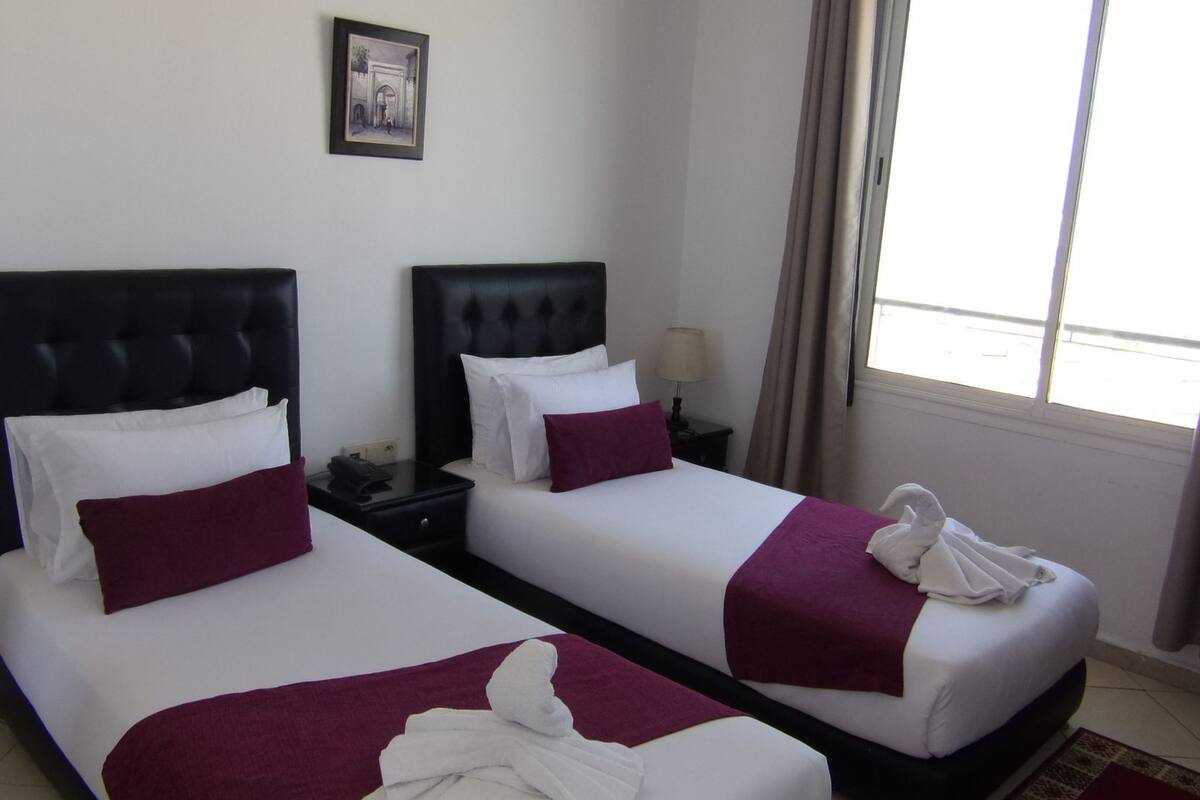 A hotel room with two beds, each with a black headboard, white bedding, and a purple throw blanket. There is a framed picture on the wall and a window with curtains.