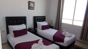 A hotel room with two beds, each with a black headboard, white bedding, and a purple throw blanket. There is a framed picture on the wall and a window with curtains.