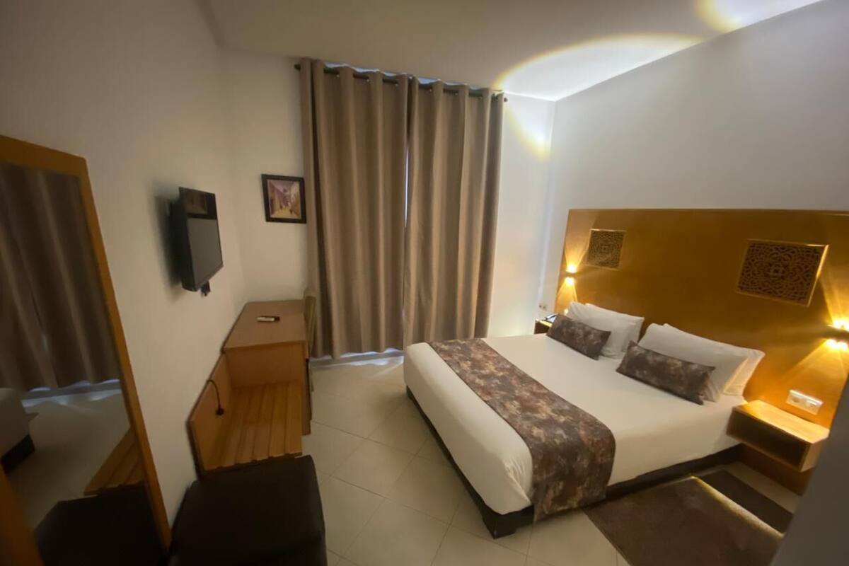A hotel room with a bed, bedside tables, a desk, a chair, a TV, and curtains.