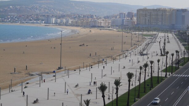 Un paisaje urbano costero con un amplio paseo marítimo, una playa y una carretera paralela al paseo.