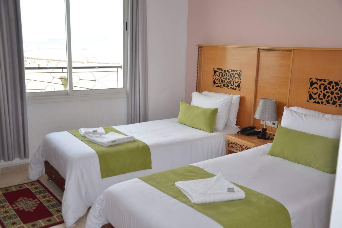 A hotel room with two beds, a window with curtains, a wooden headboard, and a telephone on the nightstand.
